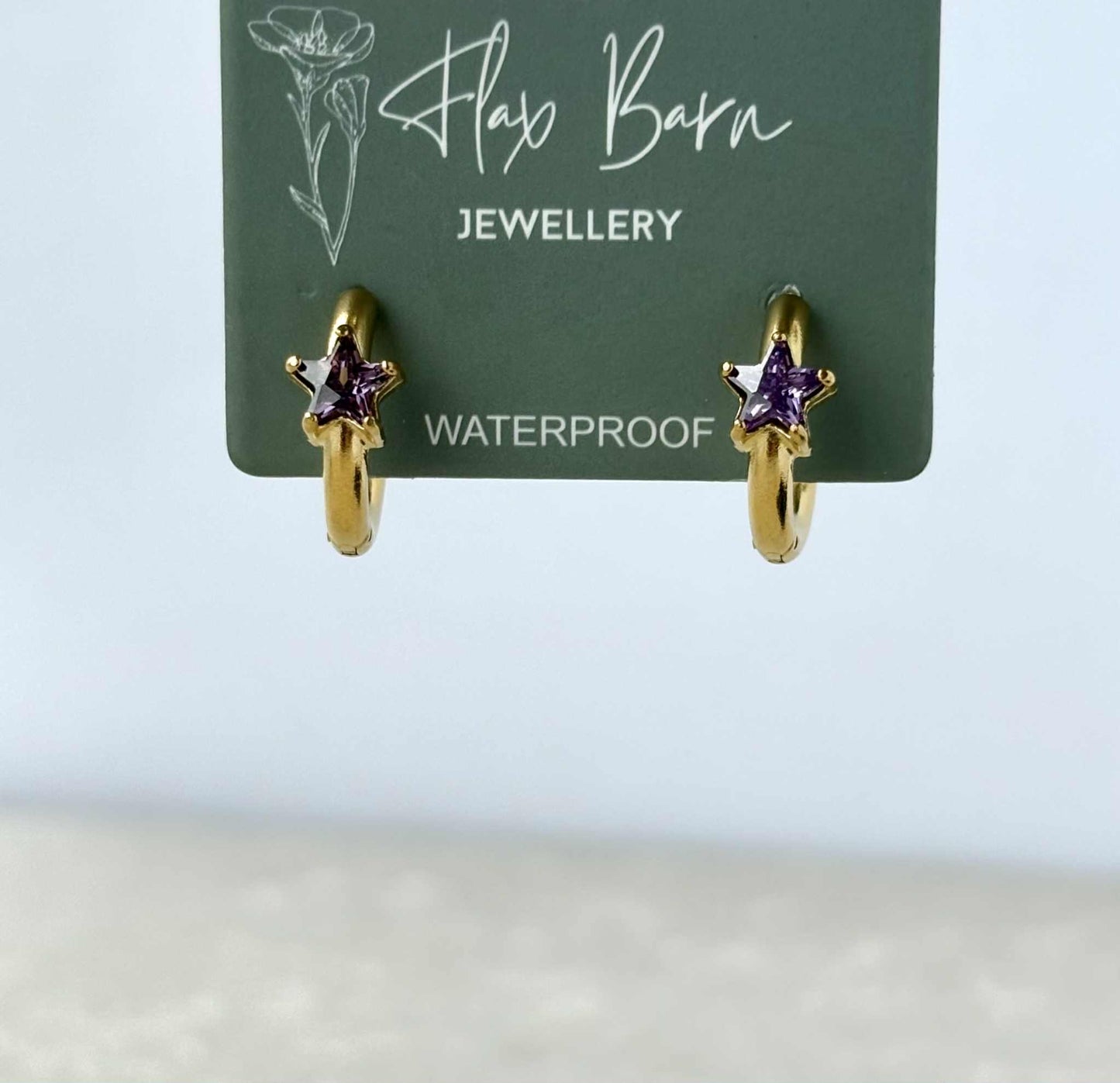 Gold hoop earrings with purple gemstones on a Flax Barn Jewellery card.