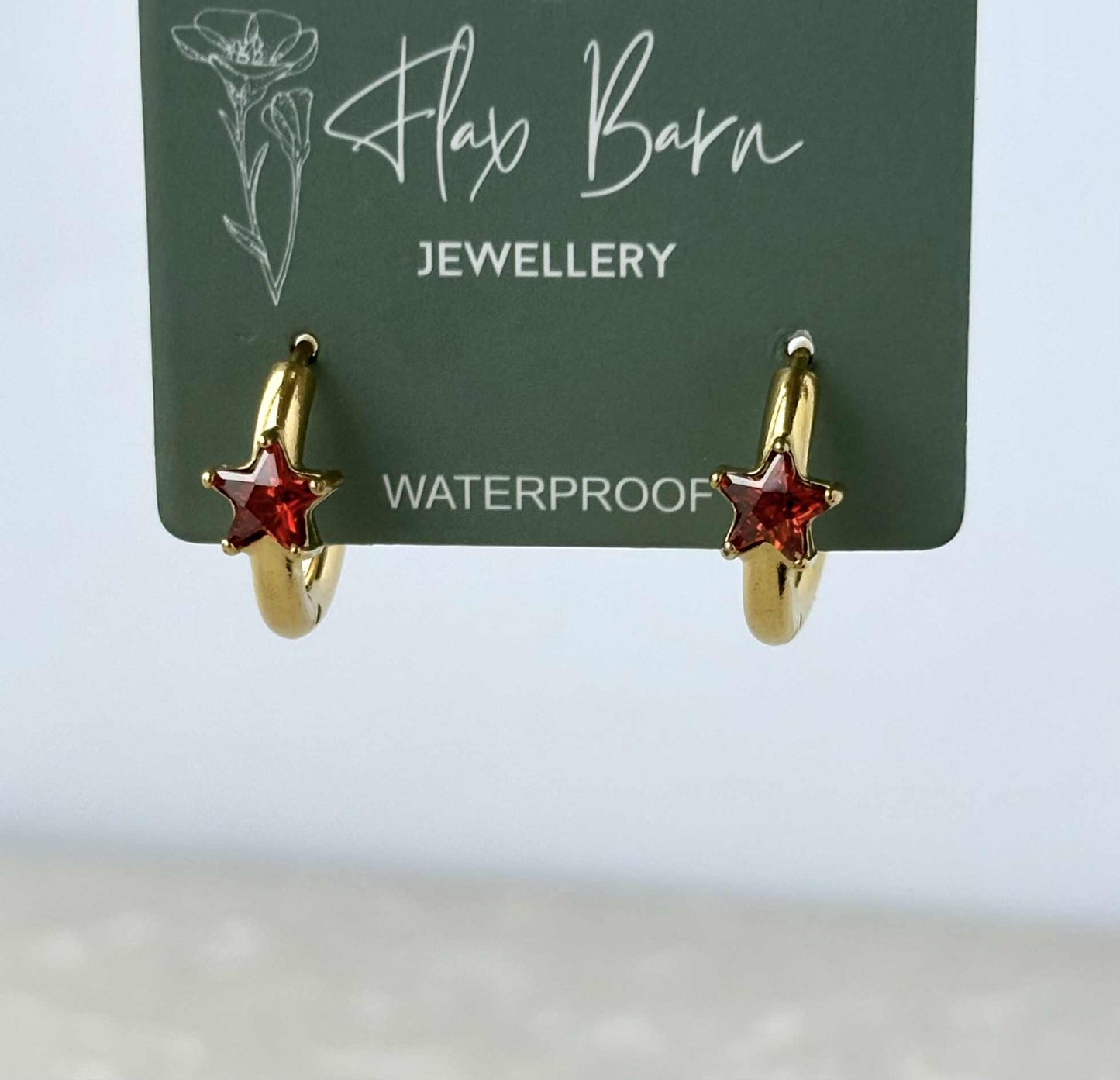 Gold hoop earrings with red star charms on a Flax Barn Jewellery card.